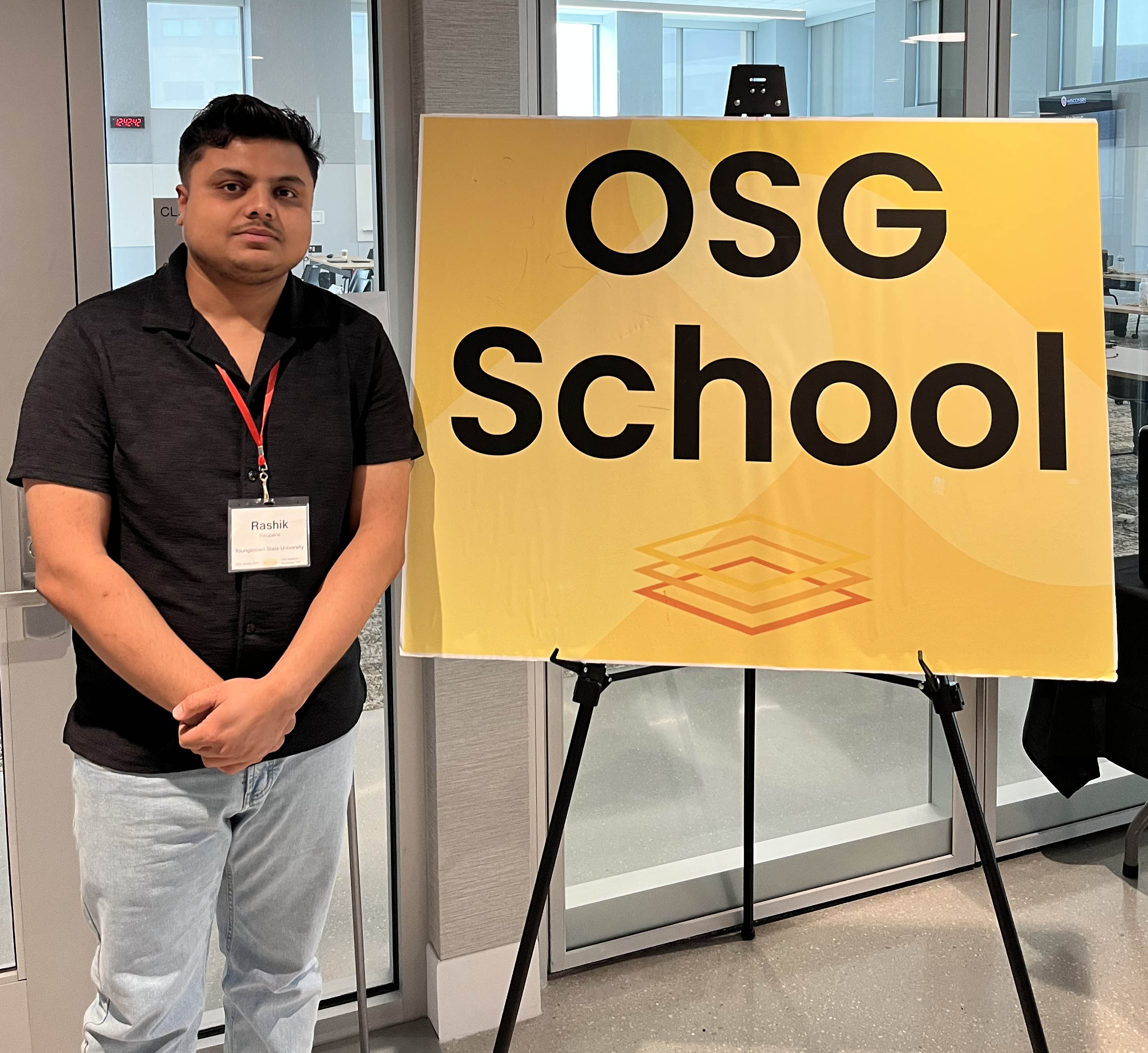 Rashik Neupane poses next to a sign that says OSG School