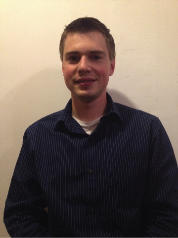A smiling male student posing against a wall and wearing a blue button down shirt